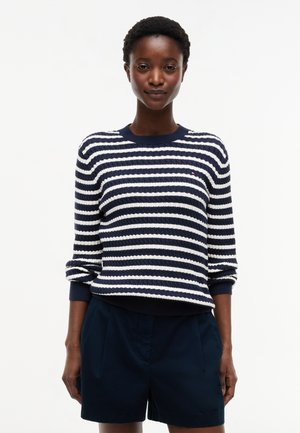 Kvinde med kort krøllet hår iført en marineblå og hvidstribet sweater og marineblå shorts, stående mod en ensfarvet hvid baggrund.