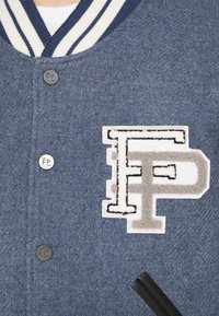 Blå ullblandning varsityjacka med en vit och marinblårandig krage, grå och vit "FP" patch och en svart tryckknapp.