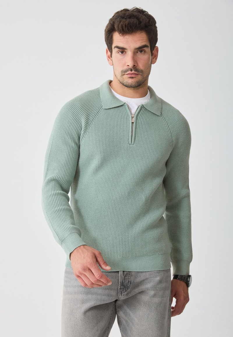 Light green ribbed pullover with half-zip collar, long sleeves, and fitted hem. Worn over a white shirt and paired with gray jeans.