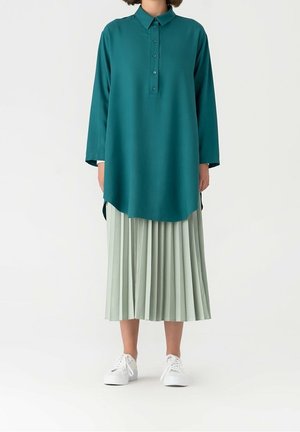 Personne portant une tunique à manches longues teal avec col, une jupe midi plissée vert clair et des baskets blanches à plateforme, debout sur fond blanc.