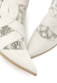Botas blancas hasta el tobillo con punta afilada, material de ante y recortes de encaje integrados en el diseño para mayor textura y contraste.