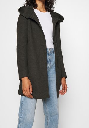 Femme portant un manteau à capuche gris foncé sur une chemise blanche et un jean bleu clair, debout devant un fond blanc uni.