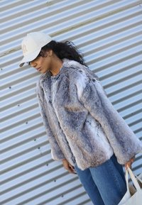 Faux fur jas in grijs en witte tinten, met een losse pasvorm, ronde halslijn en lange mouwen. Gecombineerd met een blauwe jeans en een beige pet.