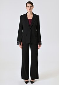 Completo nero su misura con maniche rifinite in pizzo, abbinato a un top bordeaux. Presenta un blazer aderente e pantaloni a gamba larga. Scarpe con tacco alto.