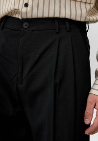 Zwarte plissébroek van gladde stof, met een knoopsluiting, zijzakken en een getailleerde pasvorm. Verticale strepen zichtbaar op het shirt.