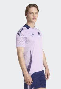 Ljuslila träningsshirt med marinblå krage och tre vertikala ränder på axlarna, med en texturerad marinblå sidopanel och logo.
