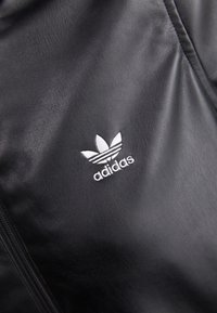 Zwarte leren jas met een opvallend witte geborduurde Adidas trefoil logo aan de linkerkant, met een zachte textuur en ritsdetails.