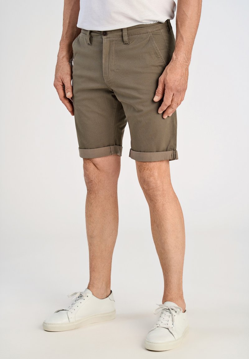 Shorts en coton vert olive avec revers roulés, deux poches avant, une fermeture par bouton, et un motif de tissu texturé. Portés avec des baskets blanches.