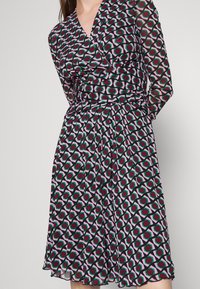 Kobieta w długiej sukience z dekoltem w kształcie litery V, z geometrycznym wzorem w kolorach fioletowym, zielonym i czarnym, stoi z rękami za plecami.