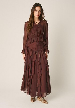 Mujer con un vestido largo, transparente y marrón oscuro con volantes, cintura fruncida y botas de tobillo con estampado floral, posando frente a un fondo liso.