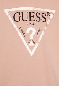 T-shirt couleur pêche arborant un logo triangulaire avec le texte "GUESS" et "U.S.A.", ainsi que d'autres détails en noir. Texture lisse.