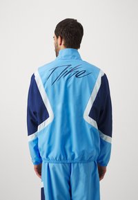 Lyseblå jakke med marineblå detaljer, der har høj krave, elastisk talje og et stort "Nike"-logo på ryggen. Glat tekstur.