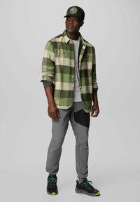 Grün-beige karierte Flanellshirt über einem grauen T-Shirt; graue Hose mit einem schwarzen Patch, schwarze Schuhe mit grünen Akzenten und eine grüne Mütze.