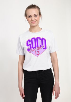 Ung kvinde iført en hvid T-shirt med lilla og pink tekst "SOCO Don't Wait! 13" og sorte bukser, stående foran en ensfarvet baggrund.