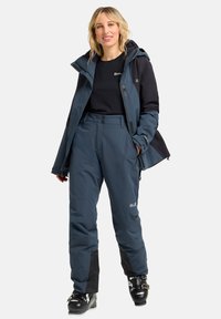 Blaue isolierte Skijacke mit Reißverschluss vorne, Kapuze und schwarzen Ärmeln. Kombiniert mit passenden blauen Skihosen und schwarzen Skischuhen.