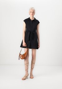 Vestido preto de camisa com mangas curtas, frente com botões e cintura atada. Modelo carrega uma bolsa de mão castanha e texturizada. Sandálias de salto alto beges com tiras.