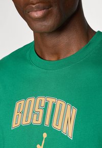 Camiseta de algodón verde con la palabra "BOSTON" en letras doradas en negrita y una silueta de baloncesto, con cuello redondo y mangas cortas.