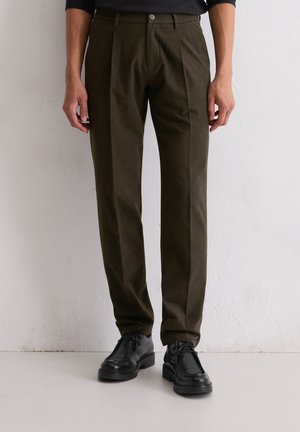 Donkergroene olijfgroene broek gemaakt van stof met een gladde textuur, met een voorplooien, knoopsluiting en zijzakken. Zwarte schoenen hieronder.