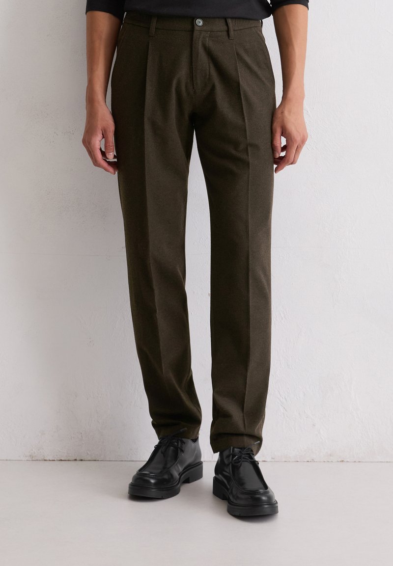 Donkergroene olijfgroene broek gemaakt van stof met een gladde textuur, met een voorplooien, knoopsluiting en zijzakken. Zwarte schoenen hieronder.