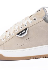 Beige suede sneaker with white laces and perforated detailing. Features a black logo accent and a brown rubber sole.