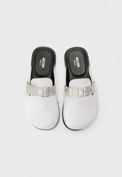 Baltie slip-on koka zābaki ar lielu sudraba "Moschino" logo uz siksnas, melnajām iebūvētajām zolēm un teksturētām melnām gumijas zolems.