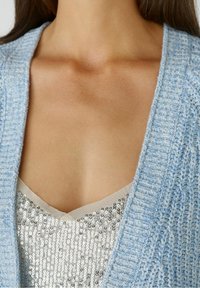 Vaaleansininen neulottu neuletakki, jossa on teksturoitu kuvio, kerroksittain hopeanvärisen paljettopaidan päällä, jossa on v-kaula-aukko.