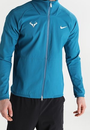 Man draagt een blauw sportjack met ritssluiting, met Nike- en stierenlogo op de borst, gecombineerd met een zwarte broek, staand tegen een effen achtergrond.