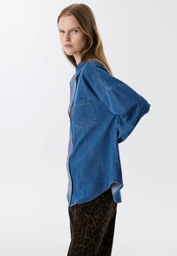 Camisa de mezclilla con un corte relajado, mangas largas y un bolsillo de parche. La tela tiene un tono azul medio con una textura suave.