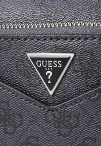Lommebok i svart teksturert syntetisk materiale med et trekantet sølvlogo med "GUESS USA" og et spørsmålstegn-designt. Glidelåsdetalj synlig.
