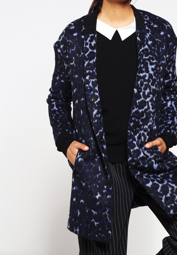 Persona che indossa un cappotto a fantasia blu e nero sopra un maglione nero con colletto bianco, mani in tasca e pantaloni a righe.