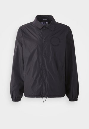 Sort letvægtsjakke med knaplukning foran, elastiske cuffe, snøre i bunden og broderet logo på brystet. Glat, let skinnende stof.