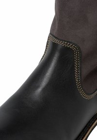 Botte en cuir noir avec une tige en suède gris foncé, des coutures contrastées, une forme arrondie et un panneau latéral en élastique pour un enfilage facile.