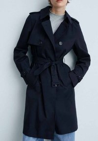 Granatowy trench coat z podwójnym zapinaniem z przodu, dużymi klapami i paskiem w talii. Posiada boczne kieszenie i paski przy mankietach. Gładkie wykończenie materiału.