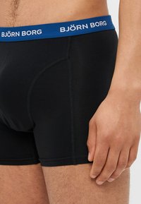 Boxer-culottes en coton noir avec une ceinture élastique bleue sur laquelle est inscrit "BJÖRN BORG" en blanc. Des coutures accentuent l'ajustement.