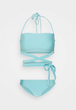 Bikini bandeau bleu clair avec des bretelles croisées ; le mannequin porte des tongs assorties et un sac en toile tissé. L'arrière-plan est d'un blanc uni.