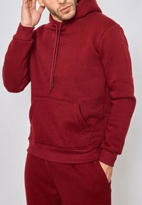 Haut à capuche bordeaux en tissu doux avec une poche avant, un capuchon à cordon ajustable et des poignets côtelés, associé à un pantalon assorti.
