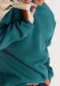 Turkusowa bluza z okrągłym dekoltem i ściągaczami na rękawach. Gładka faktura tkaniny. Beżowe paski trzymane przez wyciągniętą dłoń.