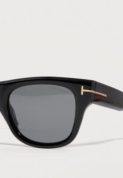 Μαύρα ορθογώνια γυαλιά ηλίου με σκοτεινούς φακούς και χρυσές διακοσμήσεις σε σχήμα Τ στους βραχίονες, τοποθετημένα σε γωνία πάνω σε λευκό φόντο.