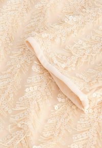 Beige tyg med paljetter ordnade i ett fjäderliknande mönster, med en mjuk textur och en rullad kant för avslutning.