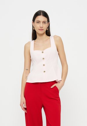 Femeie cu păr lung și închis la culoare poartă un top alb fără mâneci cu nasturi și pantaloni roșii, stând cu o mână în buzunar, pe un fundal simplu.