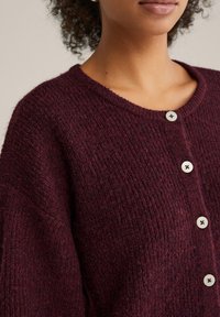 Bordo krāsas adīts kardigans ar teksturētu apdari, apaļu kakla izgriezumu un trim baltiem pogām priekšpusē. Mīksts un vidēji smags materiāls.