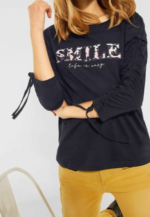 Svart långärmad skjorta med ordet "SMILE" i rosa och grått, med volangdetaljer vid ärmarna och knutna detaljer vid manschetterna.