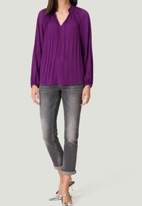 Violetti pitkähihaista paitaa, jossa on poimuttunut muotoilu olkapäillä, yhdistettynä käännettyhihaisiin harmaisiin farkkuihin ja metallisiin teräväkärkisiin korkokenkiin.