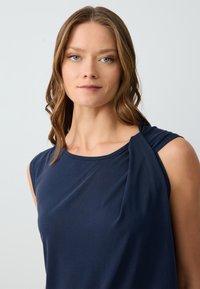 Top senza maniche blu navy in morbido tessuto drappeggiato con dettaglio arricciato vicino alla spalla, scollatura rotonda e superficie liscia.