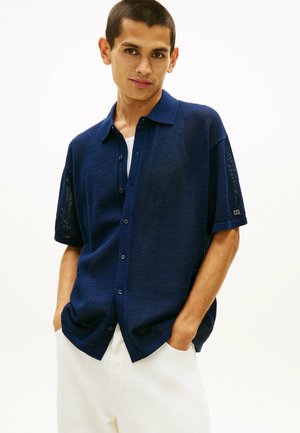 Junger Mann mit kurzem Haar, der ein marineblaues kurzärmeliges Hemd über einem weißen Oberteil und einer weißen Hose trägt, die Hände in den Taschen.