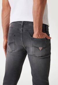 Jeans de mezclilla gris con un corte slim. Presentan un parche triangular con logo y bolsillos traseros con detalles de costura. Textura suave.