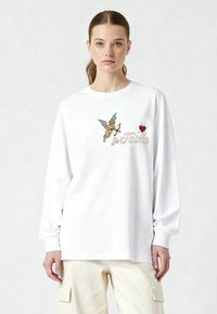 T-shirt blanc à manches longues en coton avec un motif de chérubin brodé, accompagné du texte "Je T'aime" et d'un détail cœur rouge.