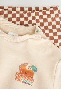 Cream baby onesie with button snaps by the neck and a smiling pumpkin pie graphic labeled "Pumpkin Pie Bake Sale" over checkered brown and cream pants.