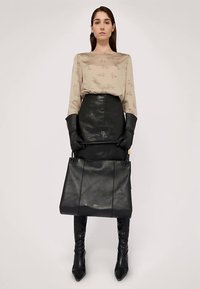 Černá kožená taška typu tote s strukturovaným designem, texturovaným povrchem a detailem loga. Modelka má na sobě béžový vzorovaný top a černé kožené rukavice.