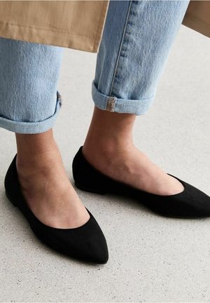 Kvinde iført lyseblå opbukket jeans og sorte spidse flade sko, der står på en lysegrå gulv.